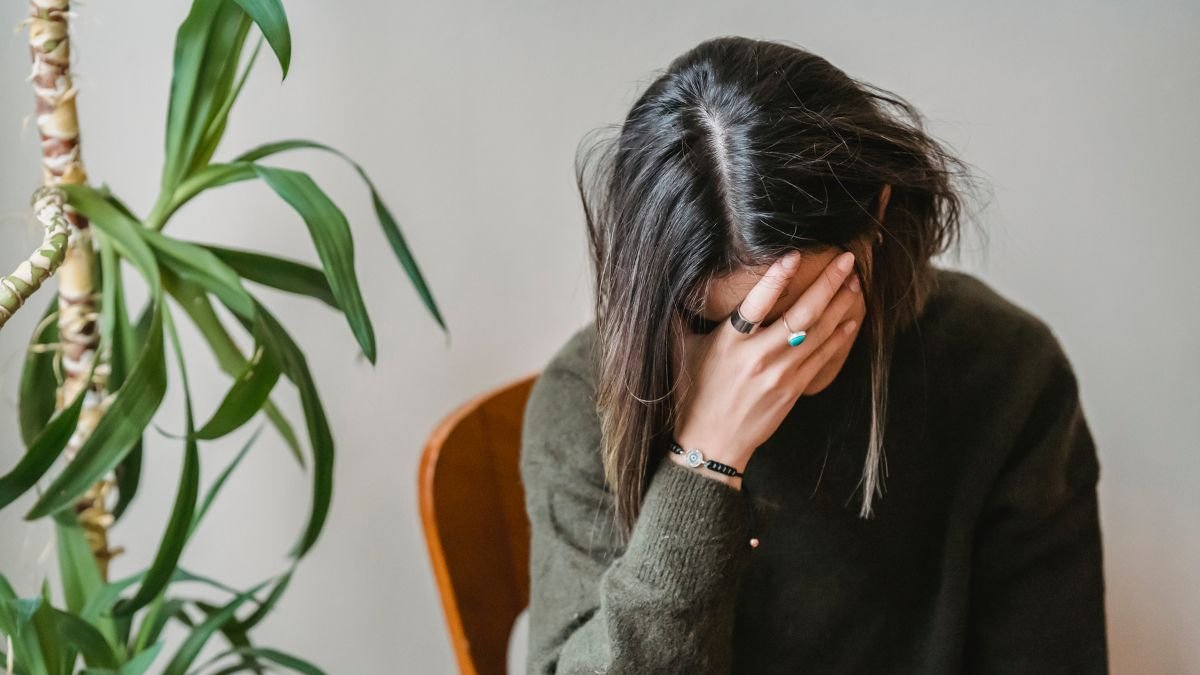 A high-resolution close-up of a woman with closed eyes and a pained expression, gently pressing her fingers to her temple, suggesting fatigue or a headache — a possible symptom of vitamin B12 deficiency.